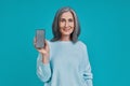 Mature beautiful woman showing smart phone and smiling while standing against blue background Royalty Free Stock Photo