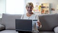 Mature blonde woman entering credit card number on laptop sitting sofa, payment