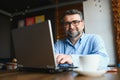 Mature businessman drinking coffee in cafe. Portrait of handsome man wearing stylish eyeglasses using laptop, looking at Royalty Free Stock Photo