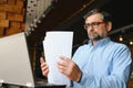 Mature businessman drinking coffee in cafe. Portrait of handsome man wearing stylish eyeglasses using laptop, looking at Royalty Free Stock Photo