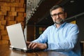 Mature businessman drinking coffee in cafe. Portrait of handsome man wearing stylish eyeglasses using laptop, looking at Royalty Free Stock Photo