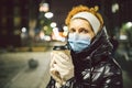 Mature Caucasian female in protective medical mask and gloves, holds takeaway coffee at night city. Senior woman walk during Royalty Free Stock Photo