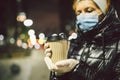 Mature Caucasian female in protective medical mask and gloves, holds takeaway coffee at night city. Senior woman walk during Royalty Free Stock Photo