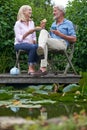 Mature Couple Enjoying Afternoon Tea And Cake Sitting On Chairs On Wooden Jetty By Lake