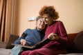 Mature daughter visits senior mother and has fun watching family photo album, sitting in embrace in living room couch. Mothers Day