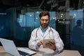 mature doctor in a medical gown is working inside clinic office, doctor is holding a tablet computer in his hands for Royalty Free Stock Photo