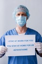 Mature doctor in medical mask and cap holding placard with i stay at work for you, you stay at home for us lettering