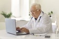 White-haired senior doctor typing on laptop keyboard sitting at desk in hospital office Royalty Free Stock Photo