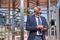 Mature, executive, gray-haired man with beard, sunglasses, jacket and tie, checking emails on his cell phone outside an office.