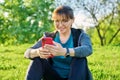 Mature female in headphones with smartphone watching video, sitting on grass Royalty Free Stock Photo