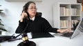 Mature female judge or lawyer or in robe gown uniform using laptop and talking on telephone at her office Royalty Free Stock Photo