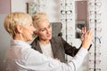 Mature female seller helping senior woman to choose prescription glasses in optician store