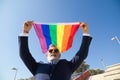 Mature gay man, executive, grey-haired, bearded, sunglasses, jacket and tie, waving the new lgbtiq+ pride flag in the wind under a