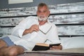 Mature, gray-haired man with beard and eyeglasses in hand, in pajamas, lying in bed looking at the camera while reading a book Royalty Free Stock Photo