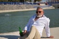 Mature, gray haired man with beard, sunglasses, white shirt and light brown pants, sitting with a bottle of beer in his hand by a Royalty Free Stock Photo