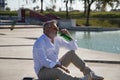 Mature, gray haired man with beard, sunglasses, white shirt and light brown pants, sitting with a bottle of beer in his mouth, Royalty Free Stock Photo