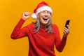 Mature gray-haired woman in santa hat holds smartphone and celebrating victory