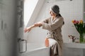 Mature grey woman applying cream while sitting in bathroom