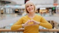 Mature happy caucasian woman standing in public place posing sincerely smiling looking at camera making heart shape with Royalty Free Stock Photo