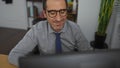 Mature hispanic man wearing glasses working in an office setting while looking at a computer screen, surrounded by indoor plants Royalty Free Stock Photo