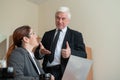 A mature male boss praises a subordinate. A woman in a suit works at a laptop at a desk. Friendly colleagues chat in the