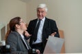 A mature male boss praises a subordinate. A woman in a suit works at a laptop at a desk. Friendly colleagues chat in the