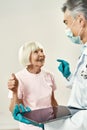 Mature male doctor in medical uniform and protective mask talking with happy senior female patient. They are showing Royalty Free Stock Photo
