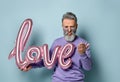 Mature male in sunglasses, purple sweater. Holding pink balloon in form of word love, kissing his palm, posing on blue background