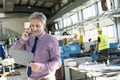 Mature male supervisor looking at clipboard while talking on mobile phone in industry