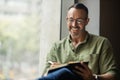 Mature man with beard, glasses writing in book sitting by sunny window