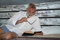 Mature man, gray-haired and bearded and biting the temple of a pair of eyeglasses, in pajamas, lying in bed reading a book quietly