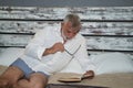 Mature man, gray-haired and bearded and biting the temple of a pair of eyeglasses, in pajamas, lying in bed reading a book quietly