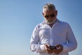 Mature man, gray haired, bearded, sunglasses, white shirt, checking social networks on his cell phone, outdoors. Concept