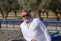 Mature man, gray-haired, bearded, sunglasses, white shirt, leaning on a railing outdoors. Concept grandfather, sugar daddy,