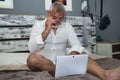 Mature man, gray-haired and bearded, wearing glasses, in pajamas, sitting on the bed with his laptop. Concept loneliness,