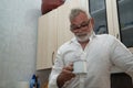 Mature man, gray-haired and bearded, wearing glasses, with white shirt and a cup of coffee in his hand, looking down with