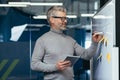 Mature man inside office near whiteboard with colored notes writing down strategy and business plan, gray haired Royalty Free Stock Photo