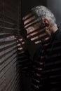 Mature man looking out of a window with blinds casting shadows Royalty Free Stock Photo