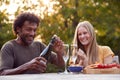 Mature Man Opening Champagne Bottle As Couple Celebrate Sitting At Table In Garden With Snacks Royalty Free Stock Photo