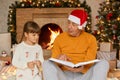 Mature man sitting on floor and holding book in hands, looking at her granddaughter and reading to child Christmas stories, little