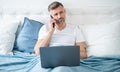 mature man speak on phone and working on laptop in bed