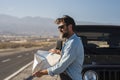 Mature man in sunglasses searching direction on location map while standing beside bonnet of jeep at highway. Handsome man reading