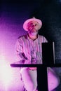 Mature man wearing a hat casually seated on a chair under purple neon lights