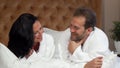 Mature married couple in bathrobes talking, lying in bed at the hotel together