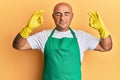Mature middle east man wearing cleaner apron and gloves relax and smiling with eyes closed doing meditation gesture with fingers