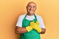 Mature middle east man wearing cleaner apron and gloves smiling with hands on chest with closed eyes and grateful gesture on face