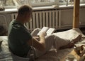 Mature modern man sitting on bed and reading book, side or back view