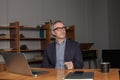 Mature pensive businessman or entrepreneur with grey hair in glasses and suit is sitting by the table