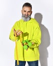 Mature positive hipster brutal bearded man in bright yellow hoodie and jeans standing with green dumbbels and tape measure