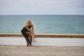 Mature, pretty, blonde woman, wearing an elegant black dress and gold heels, sitting on a stone wall, buttoning her shoes. Concept Royalty Free Stock Photo